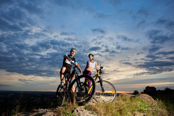 Fototapeta na wymiar Smiling young couple of cyclists in helmets sitting on mountain bicycles and looking at camera. Athletic boyfriend and beautiful girlfriend posing on rock hill against cloudy evening sky background.