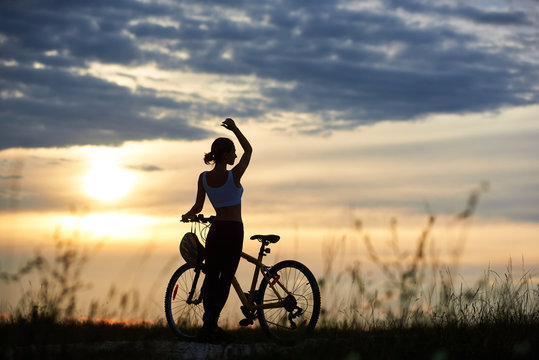 Side view of a tender girl near a bike with a raised hand upwards relaxing on the road amongst the grass on a blurred sunset background with an unusual sky with clouds and a sitting sun.