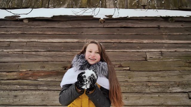 Young beautiful girl having fun blowing snow on the camera in winter clothes on the background of a wooden house in the village. Winter fun.