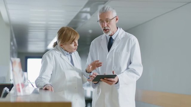 In the Hospital Two Doctors Standing at the Reception Desk, Talking and Sharing Information on Tablet Computer. Modern Hospital with Patients and Medical Personnel. 