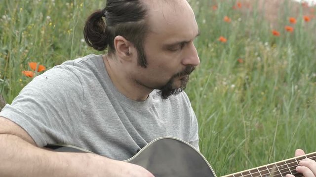 Men is playing black acoustic guitar outside in the field.