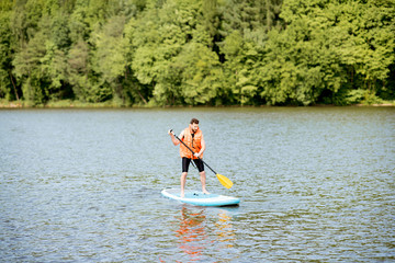 Man in life vest swimming on the standup paddleboard on the lake near the green forest