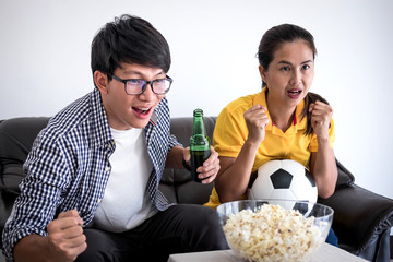 Young Asian couple love watching soccer match on tv and cheering football team, celebrating with beer and popcorn at home, sports and entertainment concept