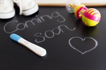 Waiting for labour, maternity concept. "Coming soon" words on black chalkboard with positive pregnancy test, white baby shoes and rattle