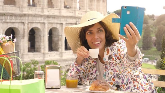 Happy young woman tourist taking selfies with smartphone at the table outside a bar restaurant in front of the Colosseum in Rome with coffee, juice and cornetto. Elegant beautiful dress with large hat