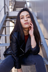 Fototapeta premium Young stylish woman in jeans and leather jacket resting on the park stairs. Attractive girl with long hair sitting on steps