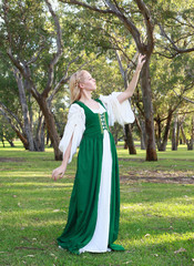 full length portrait of blonde girl wearing greensand white medieval costume, standing pose. walking through a forest.