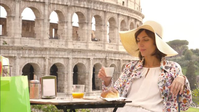 Happy young woman tourist drinking coffee and juice with cornetto at the table outside a bar restaurant in front of the Colosseum in Rome. Elegant beautiful dress with large hat and colorful shopping