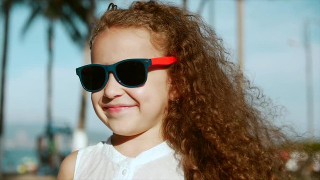 Close-up portrait of a happy cute little girl child with curly hair and red sunglasses looking into the camera