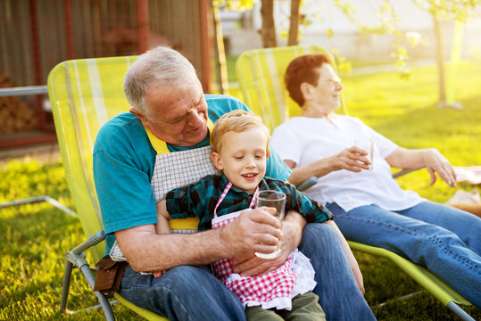 Elderly man sitting on a lazy chair outside holding his gorgeous smiling grandson and offering him water.