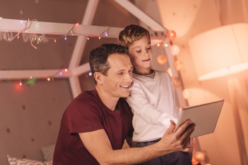 Digital technology. Nice pleasant young boy standing together with his father and pressing his finger to the tablet screen while learning to use modern technology