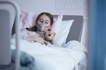 Sick girl with oxygen mask sleeping in a hospital bed with teddy bear