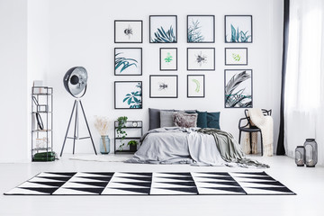 Real photo of a bed standing between a lamp and a chair in a bright bedroom interior with black and white rug and posters on a wall