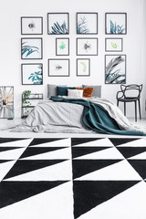 Real photo of a bed standing in a bedroom interior with black and white, triangle rug and paintings on a wall