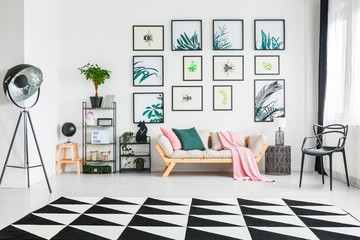 couch with cushions and a blanket standing between the shelves with ornaments and a chair in a living room interior with painting on the wall, black lamp and black and white triangle rug