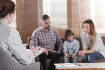 Rear view of female psychologist helping young family with a kid to solve child development problems. Family sitting on a sofa in the blurred background