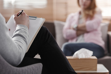 Back view of psychotherapist writing notes, assessing patient's health and giving diagnosis to a woman sitting on a couch in the blurred background during counseling session