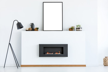 Black lamp next to fireplace in white scandi living room interior with mockup of poster. Real photo