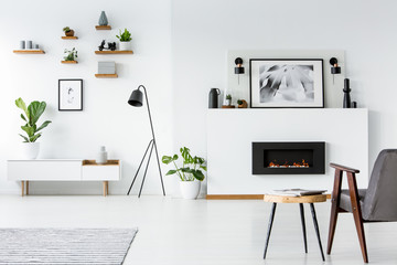 Wooden table next to grey armchair in white interior with poster above fireplace. Real photo
