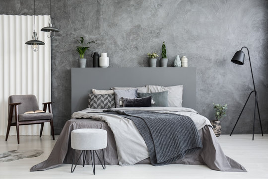 Grey stool in front of bed with blankets and cushions in dark hotel bedroom interior. Real photo