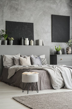 Book on stool next to bed in grey bedroom interior with black posters on dark wall. Real photo