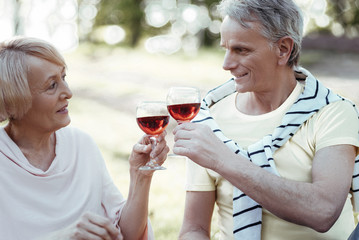 Cheers. Confident man keeping smile on his face and looking at his woman while touching glasses with her