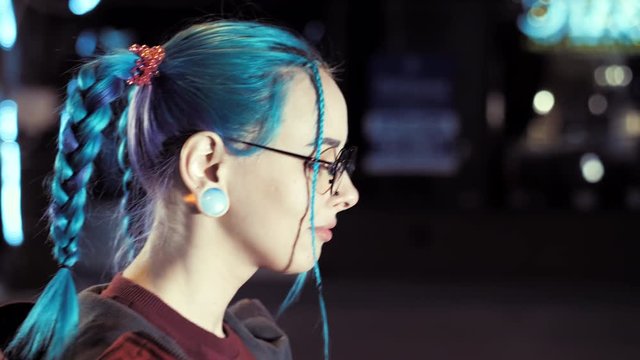 Hipster girl with blue dyed hair walking on the night city illuminated street. Portrait of teenager with piercing in nose, ears tunnels and unusual braids hairstyle. Slow motion.