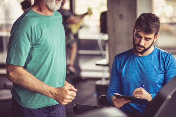 Senior man jogging on treadmill, personal trainer writing on file. Close up.