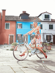 Beautiful smiling woman riding white bike near colorful houses in Burano Island, Venice, Italy. Woman dressed in romantic colorful dress, headband and red heels