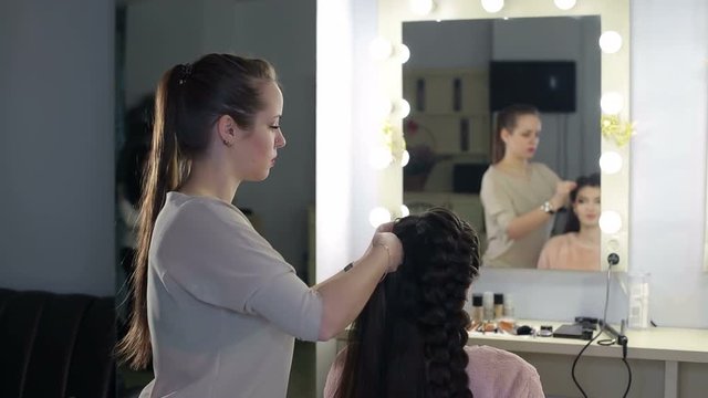 Hairdresser making big braid for client in salon. A woman hairdresser plaits a braid girl with long dark hair in front of a mirror. Weaving braids in beauty salon.
