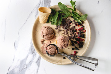 Homemade chocolate raspberry ice cream balls served with frozen berries, mint, chopped dark chocolate and metal spoon in ceramic plate over white marble background. Top view, space.