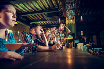 Upset male and female friends watching sport game or football match and drinking beer at bar or pub.