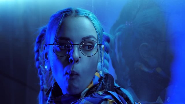 Young pretty girl with unusual hairstyle near glowing blue neon light of the city at night. Dyed blue hair in braids. Pensive hipster teenager in glasses chews cud