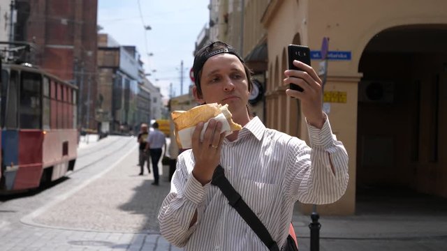 Young man stay in city public place use mobile phone and eat a big sandwich taking selfie