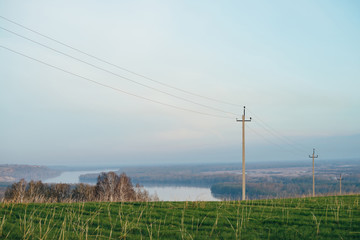 Fototapeta premium Atmospheric landscape with power lines in green field on background of river under blue sky. Electric pillars with copy space. Wires of high voltage above ground. Electricity industry.