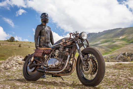 Biker with black leather suit and mask stay with his  custom special rat motorbike in a desolated mountain landscape. Post apocalyptic concept