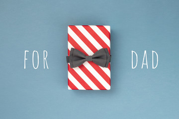 One gift box wrapped in red striped paper and tied with the grey bow tie on blue-gray background. The idea of gift design for a DAD. Holiday concept.