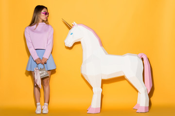 young asian female model in sunglasses holding handbag and decorative unicorn on yellow background © LIGHTFIELD STUDIOS