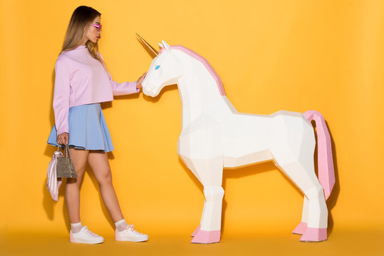 side view of asian female model in sunglasses holding handbag and touching decorative unicorn on yellow background