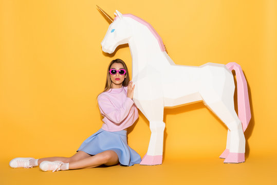 young female model in sunglasses sitting on floor and embracing decorative unicorn on yellow background
