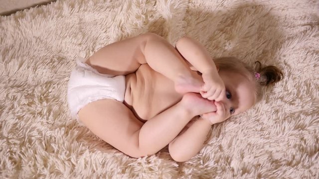Little cute toddler baby girl lie on bed bite and suck her foot toes playing with leg