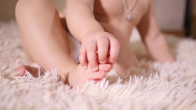 Little baby girl toddler play with her leg touch and hold her foot toe sitting on a bed