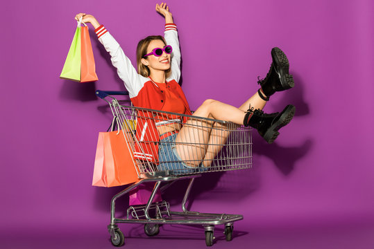beautiful happy young woman in sunglasses holding paper bags and sitting in shopping trolley on violet