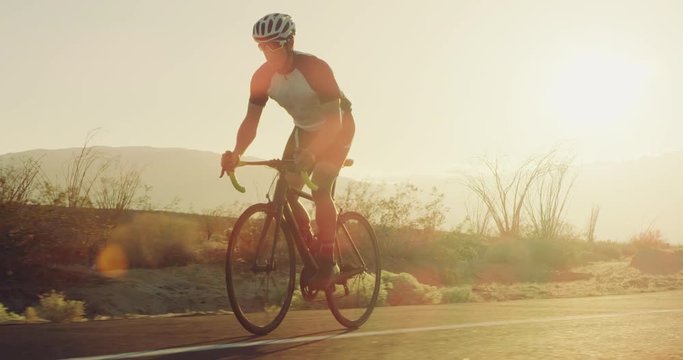 Slow motion young man cycling on road bike outside on desert road at sunset with lens flare 