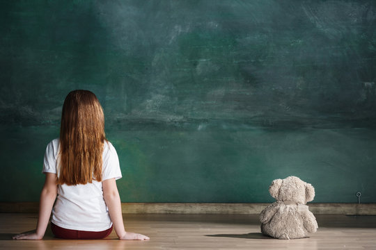 Little girl with teddy bear sitting on floor in empty room. Autism concept