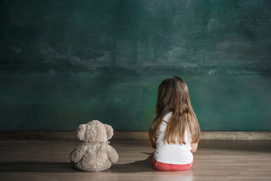 Little girl with teddy bear sitting on floor in empty room. Autism concept - Powered by Adobe