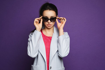 Beautiful and fashionable brunette model girl in red dress and coat adjusting her sunglasses and posing at violet background