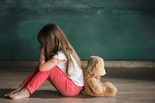 Little girl with teddy bear sitting on floor in empty room. Autism concept