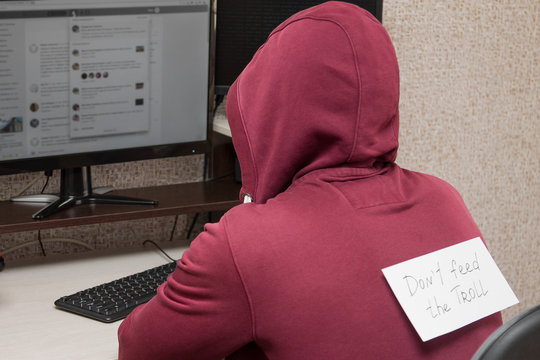 Internet troll sitting at the computer. Young man with an inscription on his back "Don't feed the Troll" writing nasty things on the forum
