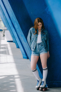 stylish woman in denim clothes, high socks and retro roller skates leaning on blue wall